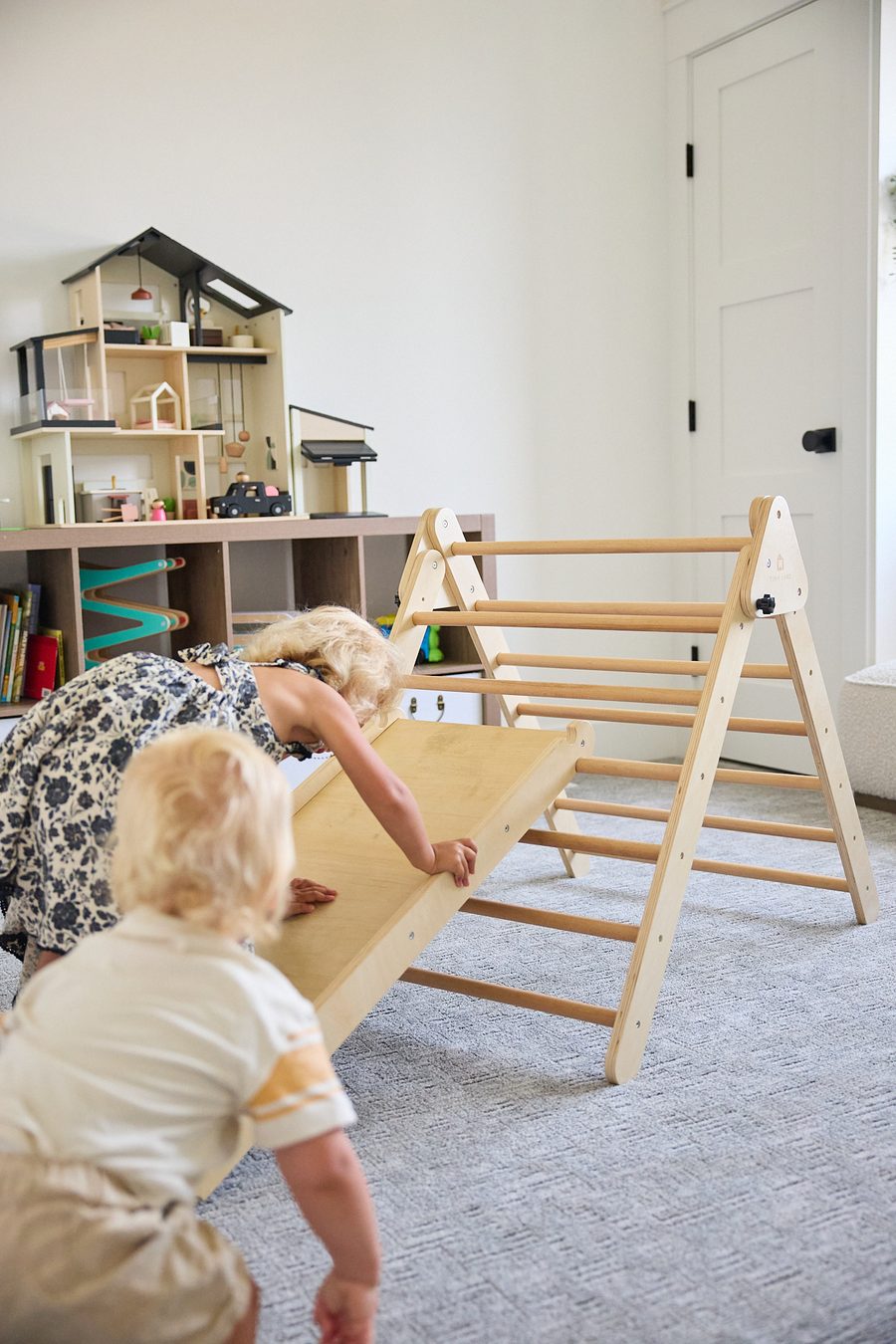 Two toddlers climbing and sliding down a wooden Pikler triangle with slide in a small living room playroom.