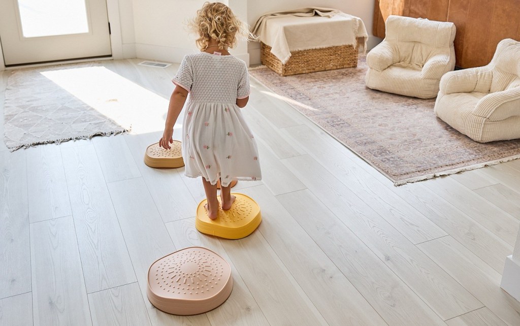 A toddler in a white dress balancing on pink, yellow, and terracotta stepping stones arranged across a bright living room floor.